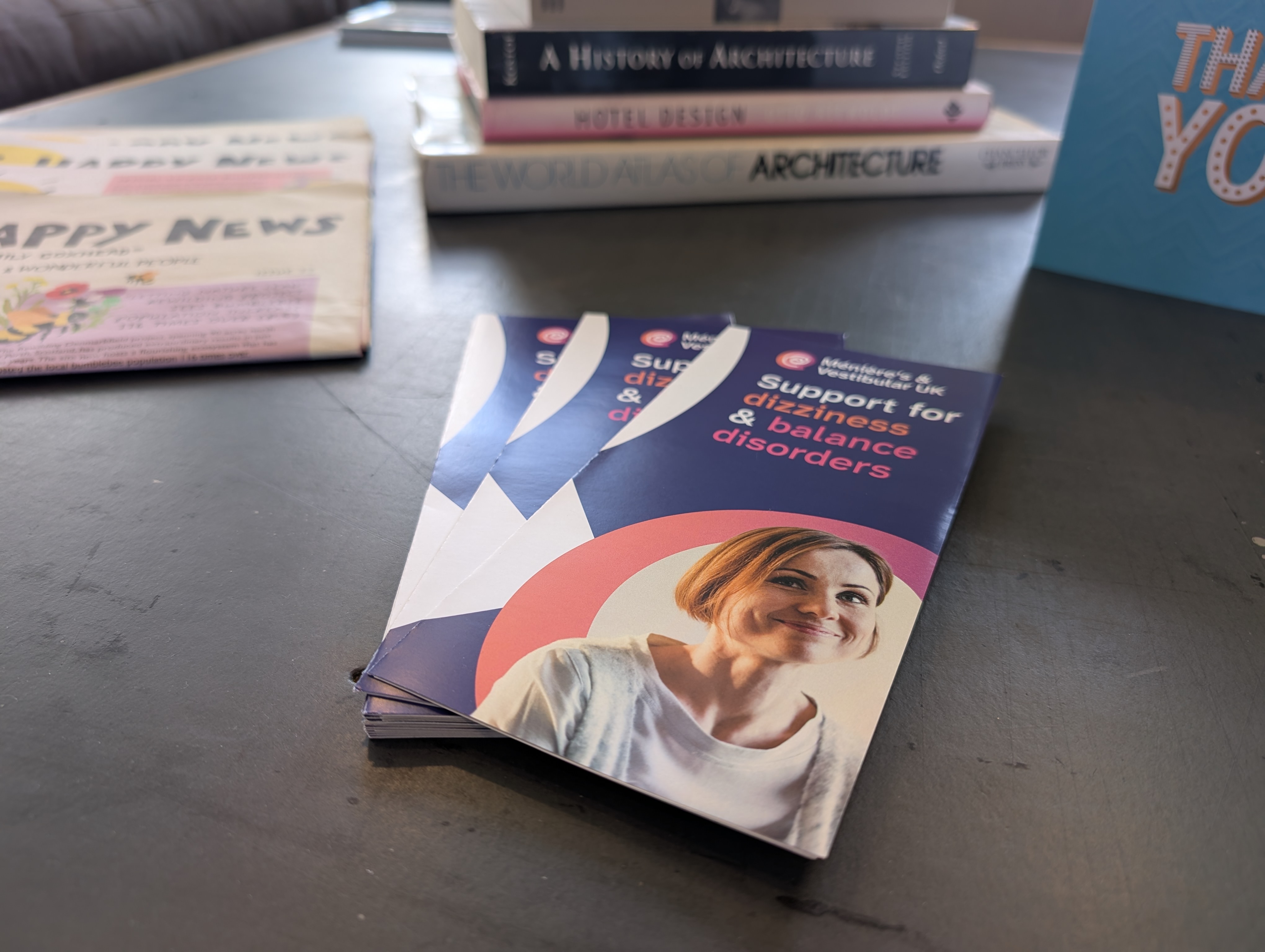 Photo of Ménière’s & Vestibular UK leaflets on our coffee table with Happy News newspapers and architectural books in the background.