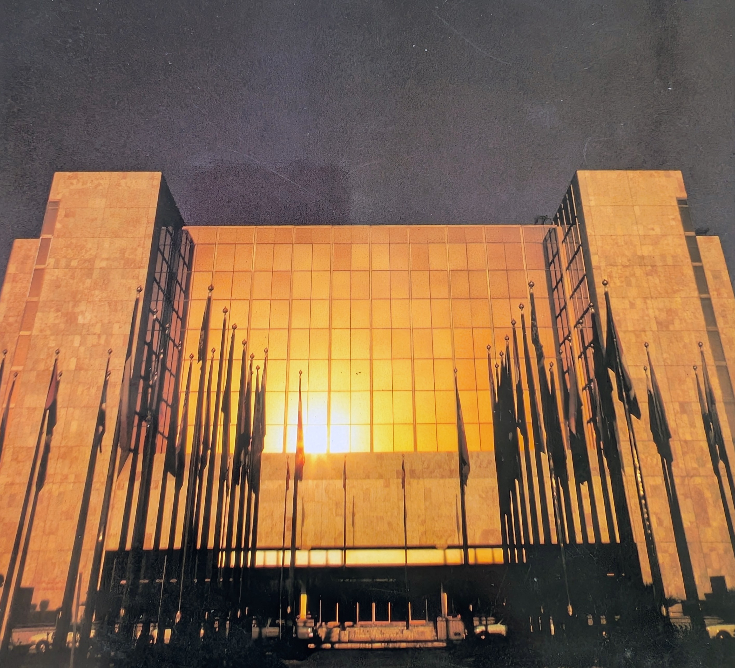 The sun reflecting a golden light off the front mirrored facade. There are lots of tall flags at the front of the hotel.