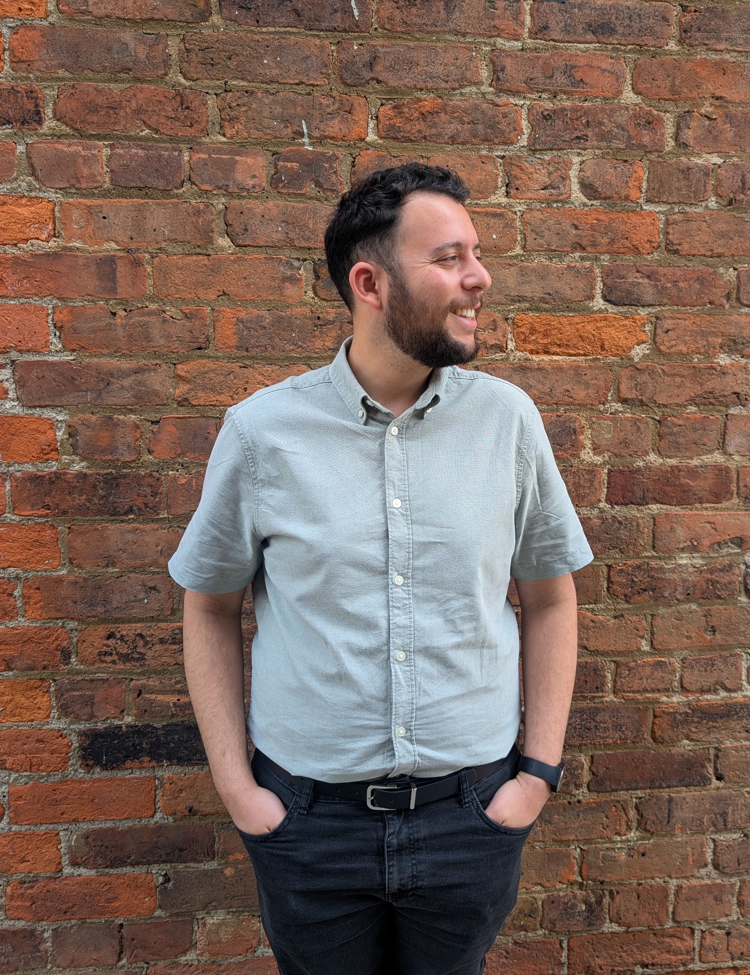 Sunny standing outside in front of a red brick wall. He’s wearing a short-sleeved sage green shirt and smiling while looking to the right of the camera.