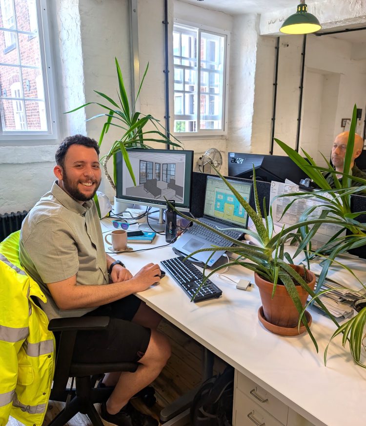 Sunny sat at his desk which is covered in plants and has plans on his screen. He’s smiling at camera.