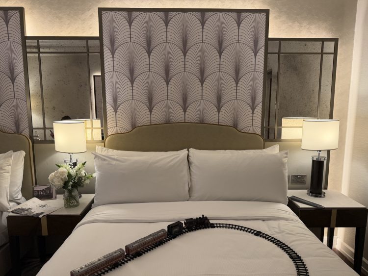 A stylish guest bedroom with a toy train track on the bed, lamps either side and a floral arrangement.