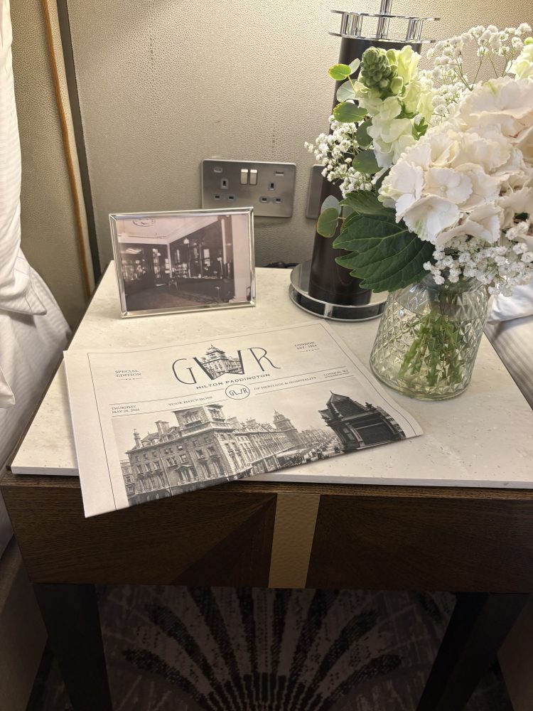 A photo of the bedside table featuring a newspaper style publication about the hotel, a black and white framed photo and white floral arrangement.