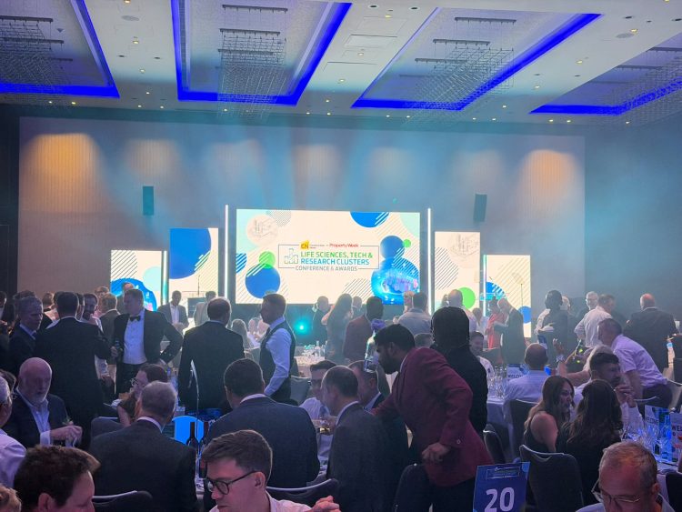 Picture of the awards dinner. Lots of people are sat round table and there is a bright screen with the awards logo at the front of the room.