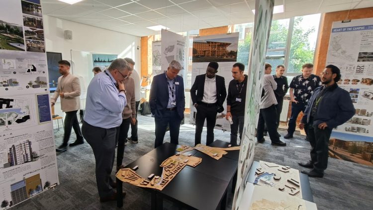 A group of people at the degree show gathered round a 3D model. Display boards are on tall panels and other people are in the background viewing the exhibition.