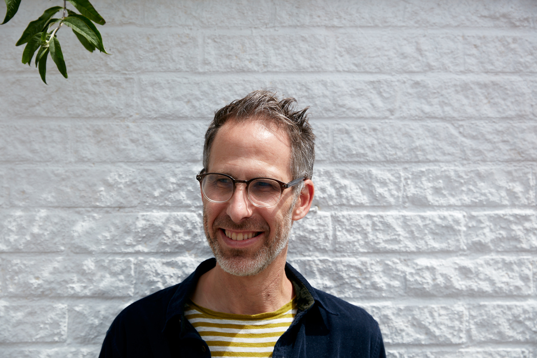 Photo of Nik Clarke stood outside in front of a white painted brick wall. He's looking to the left of the camera and smiling.