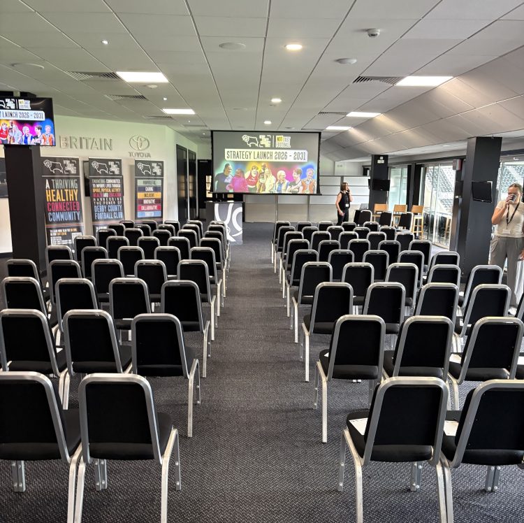 A photograph of an empty room, set up ready for a presentation, with a large screen at the front of the room.