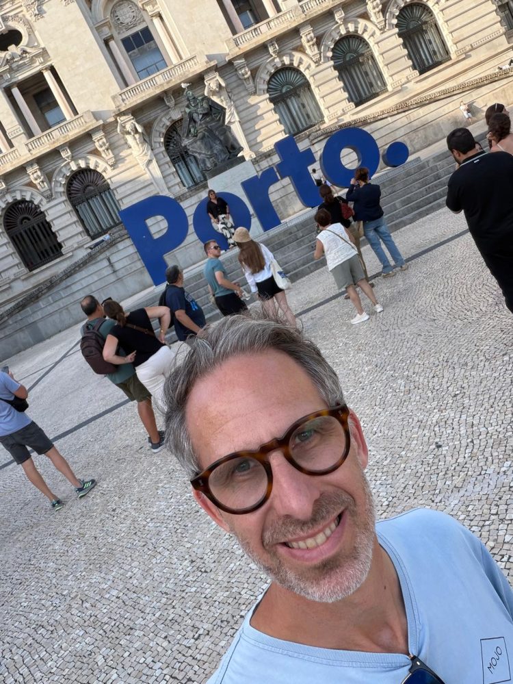 Selfie of Nik Clarke smiling at the camera with large 'Porto' 3D letters in blue behind him. He's stood in a cobbled square with an ornate stone building behind him.