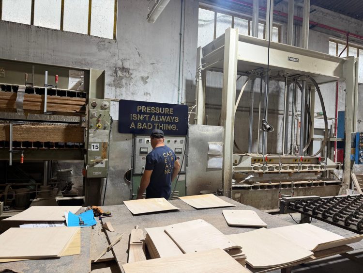 Photo of a man with his back to the camera in a production facility with machinery around him. There are pieces of wood in the foreground and a sign on the back wall which says 'pressure isn't always a bad thing'.